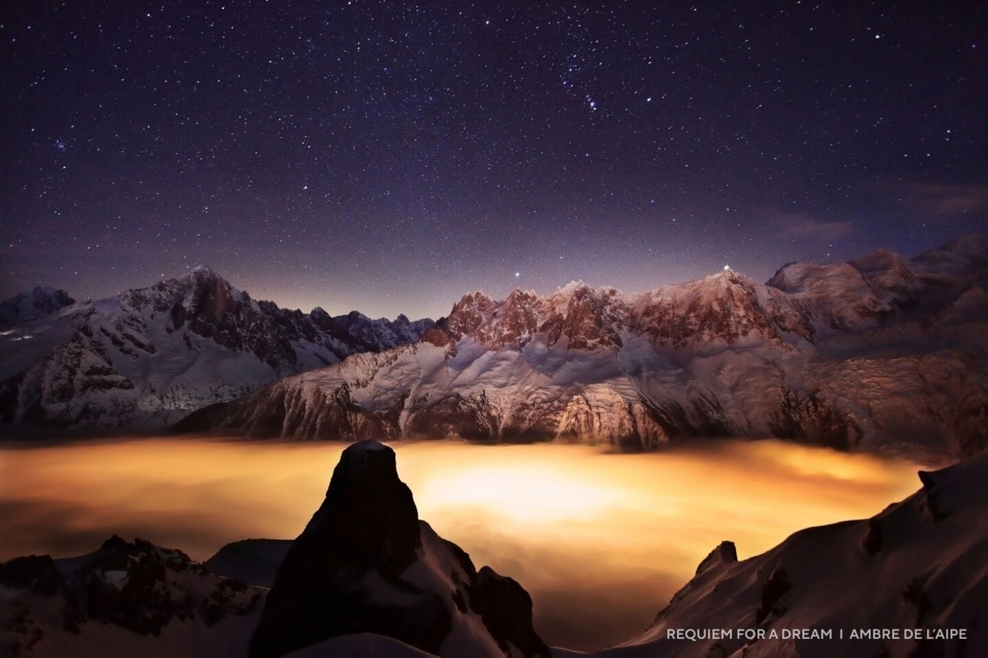 Snow-covered mountain range under the 2025 night sky, with countless stars above and a glowing orange mist or fog settled in the valley below.