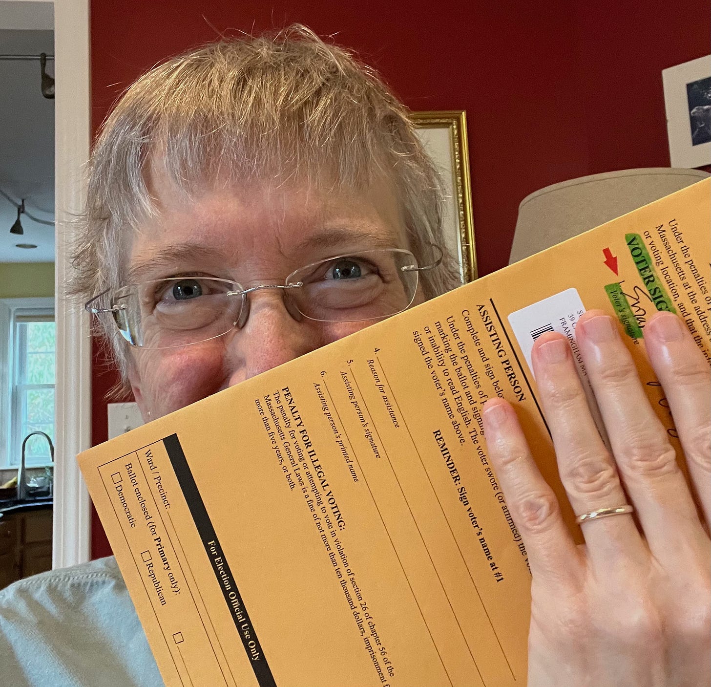 Suz, a 60-something woman with short blond hair and glasses peeks over the edge of the ballot that's ready for the dropbox!