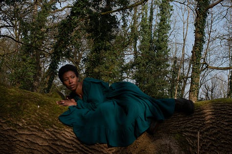 A Black woman with short afro hair wears a long, teal dress. She is reclining on moss covered fallen tree in shadow and sunlight against a backdrop of blue sky and ivy wrapped tree trunks.