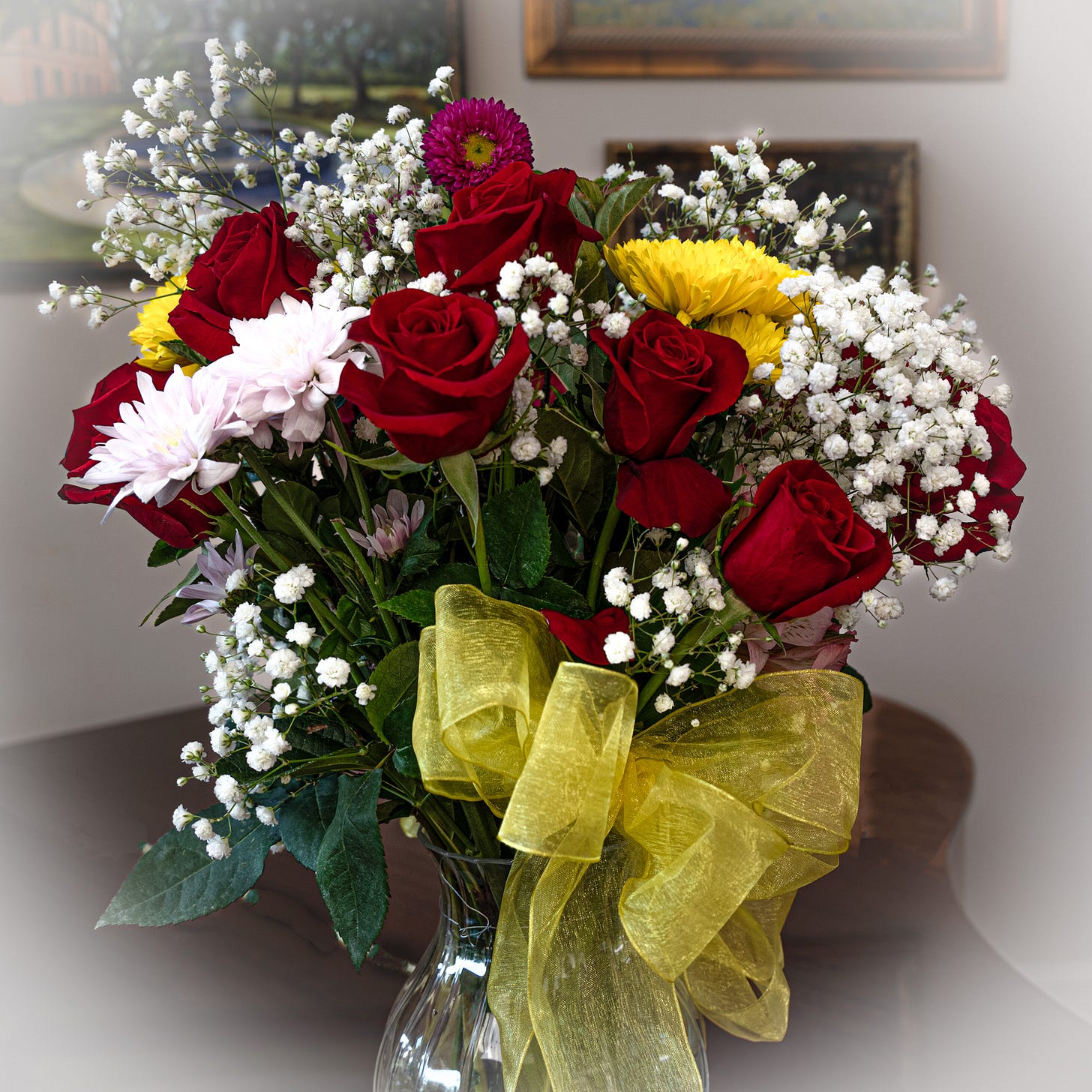 A bouquet of red roses, baby’s breath, zinnias, and yellow daisies with a yellow ribbon around the vase