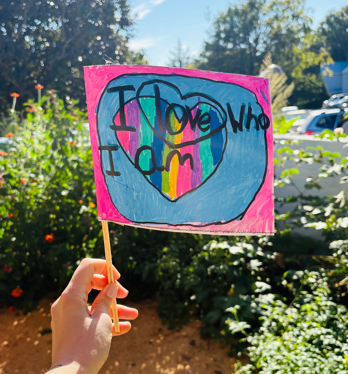 Shady holding a mini flag that says I love who I am with hearts on it. Background is a garden.