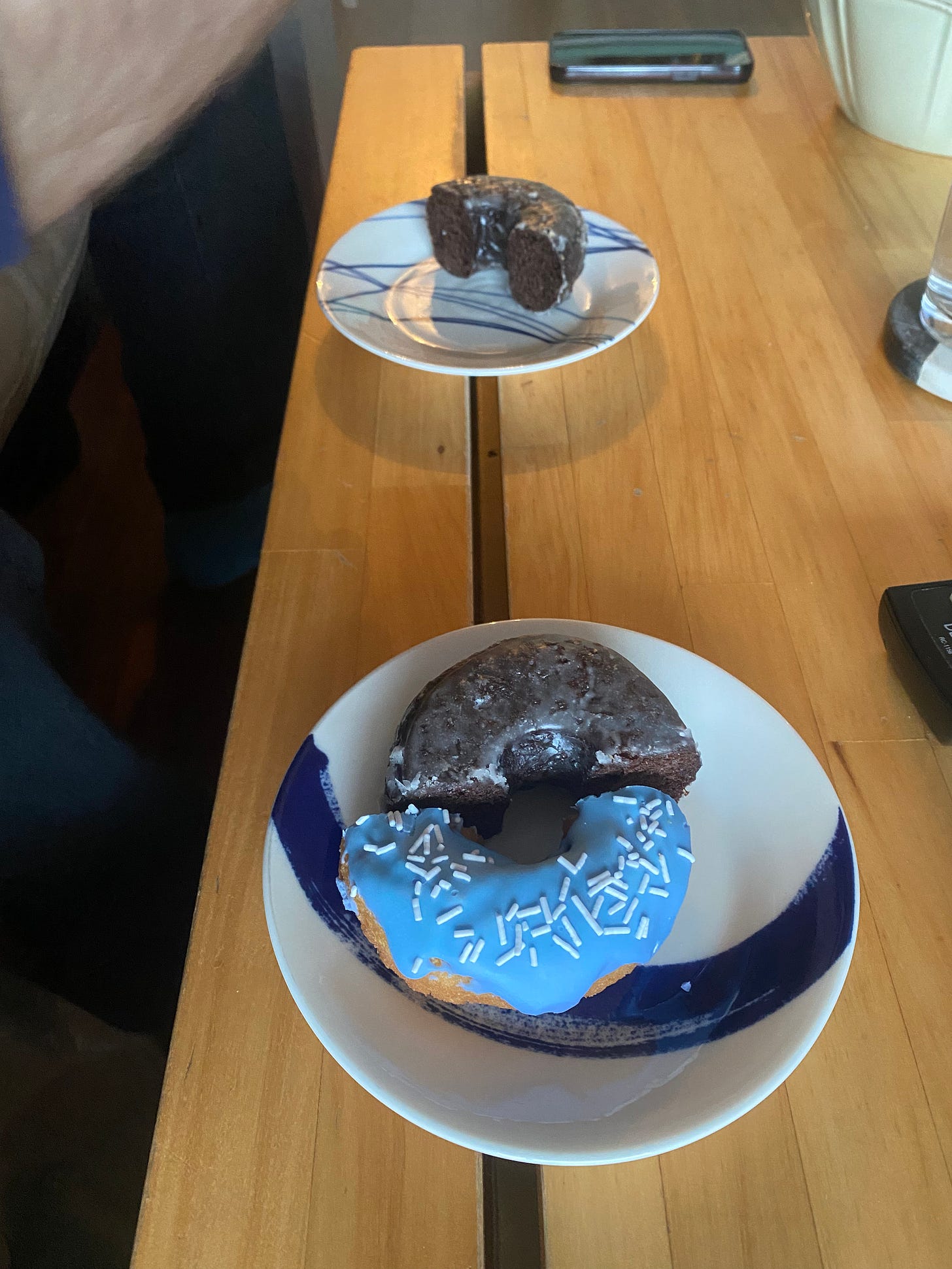two small plates with donut halves on them. The nearer of the two has a half of a chocolate glaze next to something with blue frosting and white sprinkles, and the one in the background has only the chocolate.