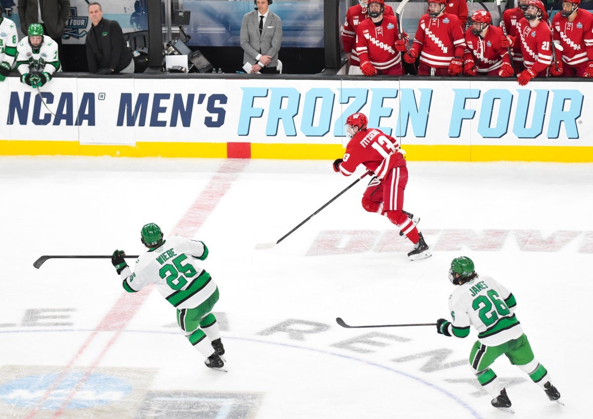 dasher board signs reading "NCAA men's Frozen Four" provide a backdrop while players chase a puck in the neutral zone
