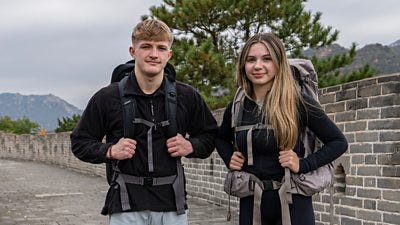 A man and woman wearing backpacks stand on the Great Wall of China smiling. Mountains are visible in the background.