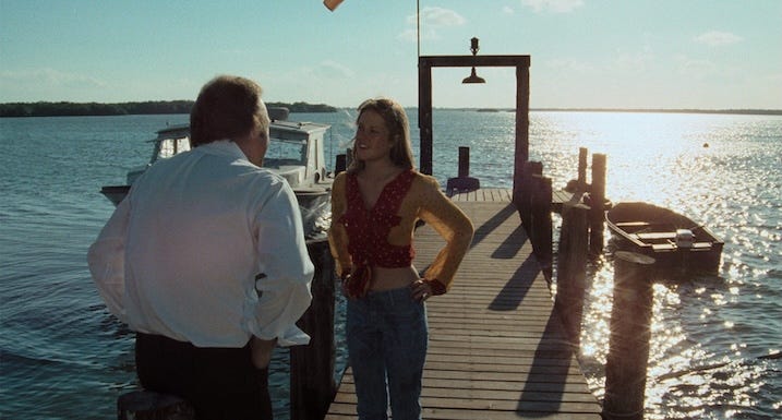Melanie Griffith stands on a sunlit dock with hands on hips, speaking to a man in a white shirt as boats shimmer in the background.