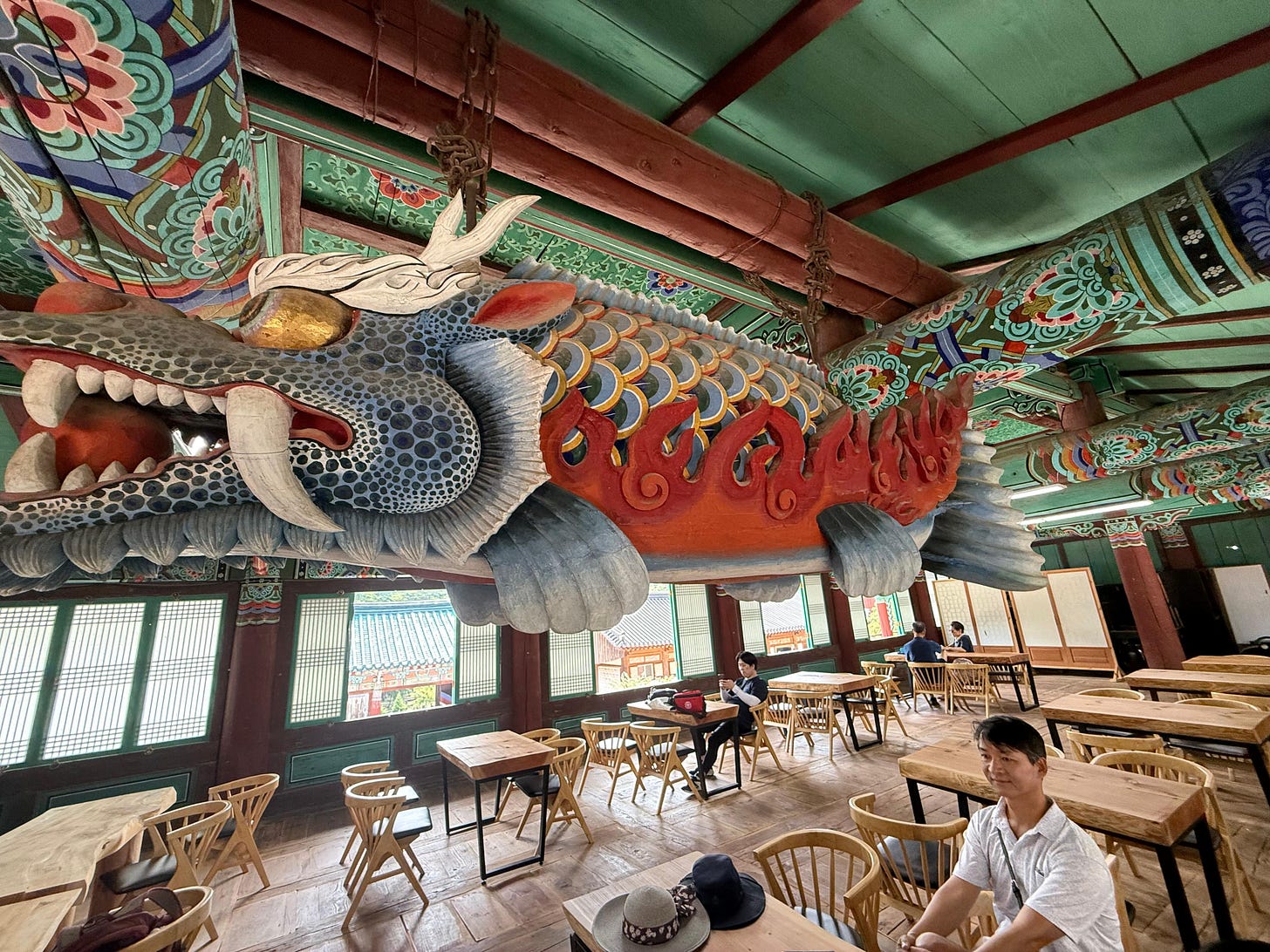 A light-filled Korean tea house filled with wooded tables and chairs and surrounded by big windows. On the ceiling, massive majestic wooden dragon weaves through the air with a man wearing a white shirt sitting underneath. A light-filled Korean tea house filled with wooded tables and chairs and surrounded by big windows. On the ceiling, massive majestic wooden dragon weaves through the air with a man wearing a white shirt sitting underneath.