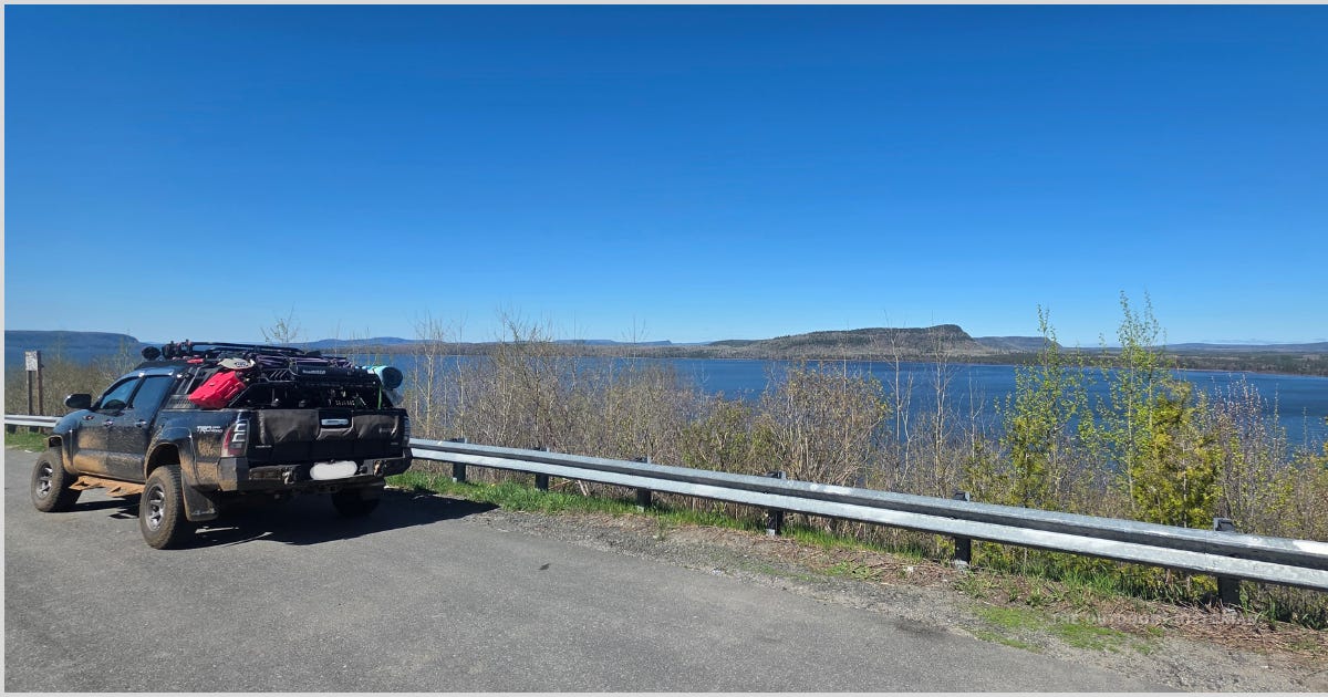 A Toyota Tacoma loaded with camping gear is parked at a scenic lookout along Highway 17 overlooking Lake Superior. The truck is viewed from behind, with blue water and a clear sky in the background.