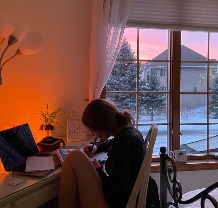 This may contain: a woman sitting at a desk with a laptop computer and writing on a notepad