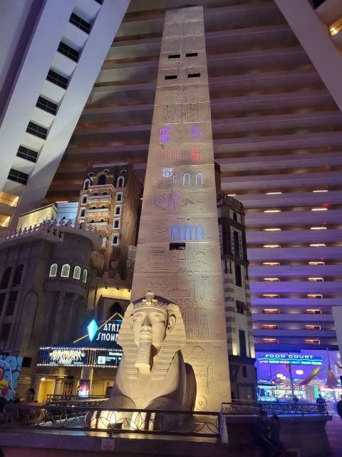 Num ambiente interno cujo teto parece lembrar uma pirâmide, um obelisco egípcio está no meio, com símbolos que imitam hieróglifos e uma pequena esfinge. Ao redor, outras construções do hotel.