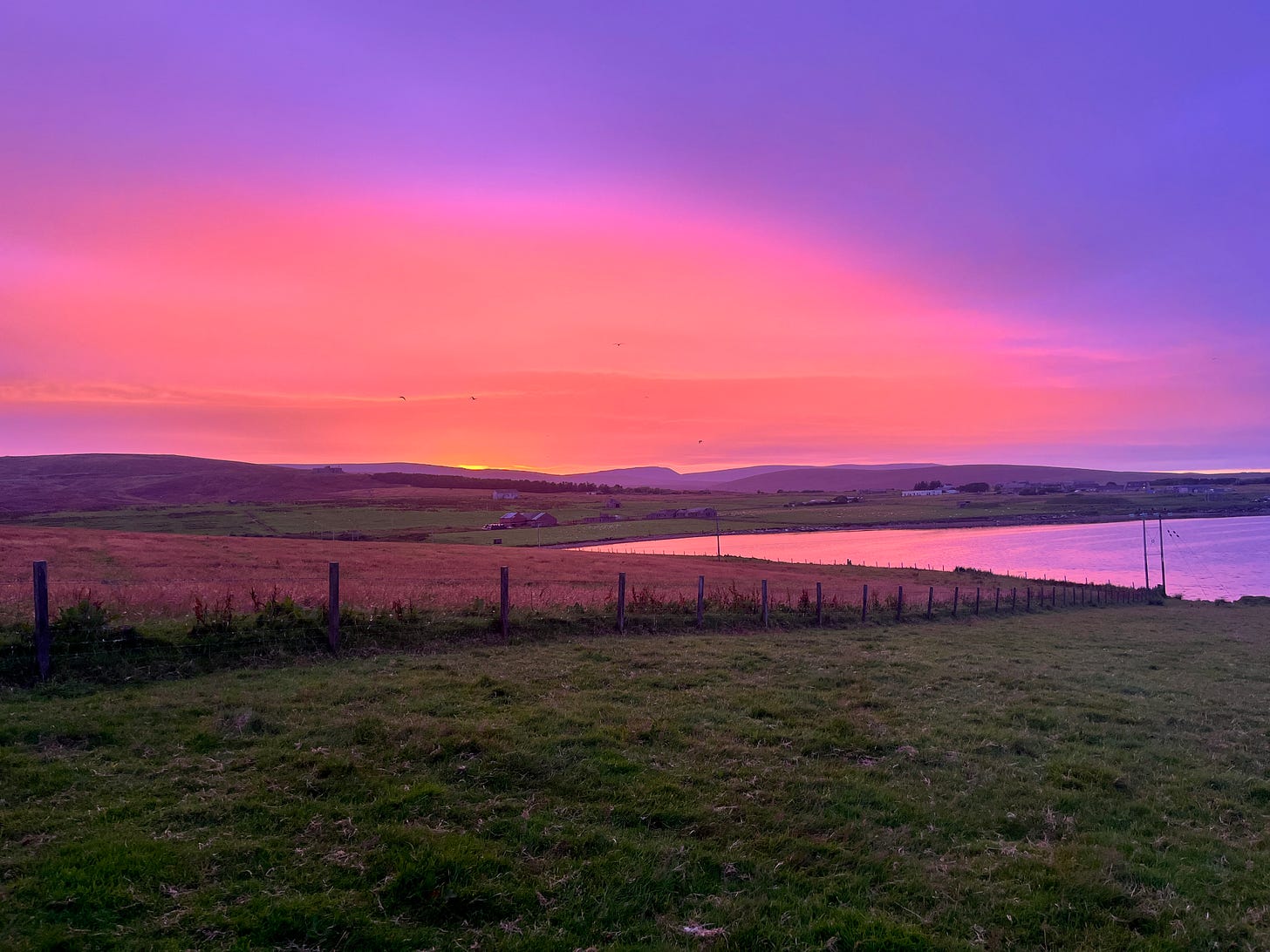 Sunset looking across grass, over a small inlet & onto hilss in the background. Colours of purple, pink & orange
