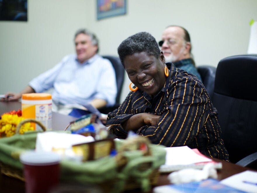 A Black woman is at a conference table, with a big smile.