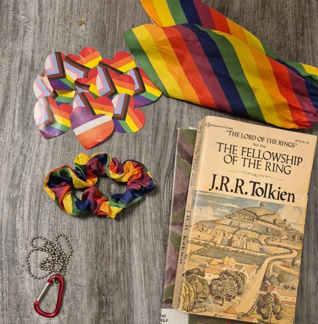 A nightstand shows a pile of pride heart stickers next to a pride scrunchie, pride bow, a carabiner on a necklace chain, and a copy of Lord of the Rings. 