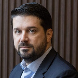 A imagem mostra um homem de pele clara, com barba e cabelos escuros curtos, olhando diretamente para a câmera com expressão séria. Ele veste blazer escuro e camisa social azul clara. Ao fundo, há uma parede com painéis verticais de madeira em tom marrom, em ambiente interno e profissional.