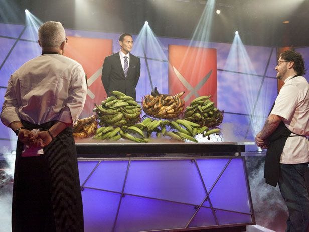 an image from Iron Chef America. Two chefs stand before a giant table laden with all manner of plantains. Dramatic host personality Mark Dacascos stands on a dais and looks down on the chefs.