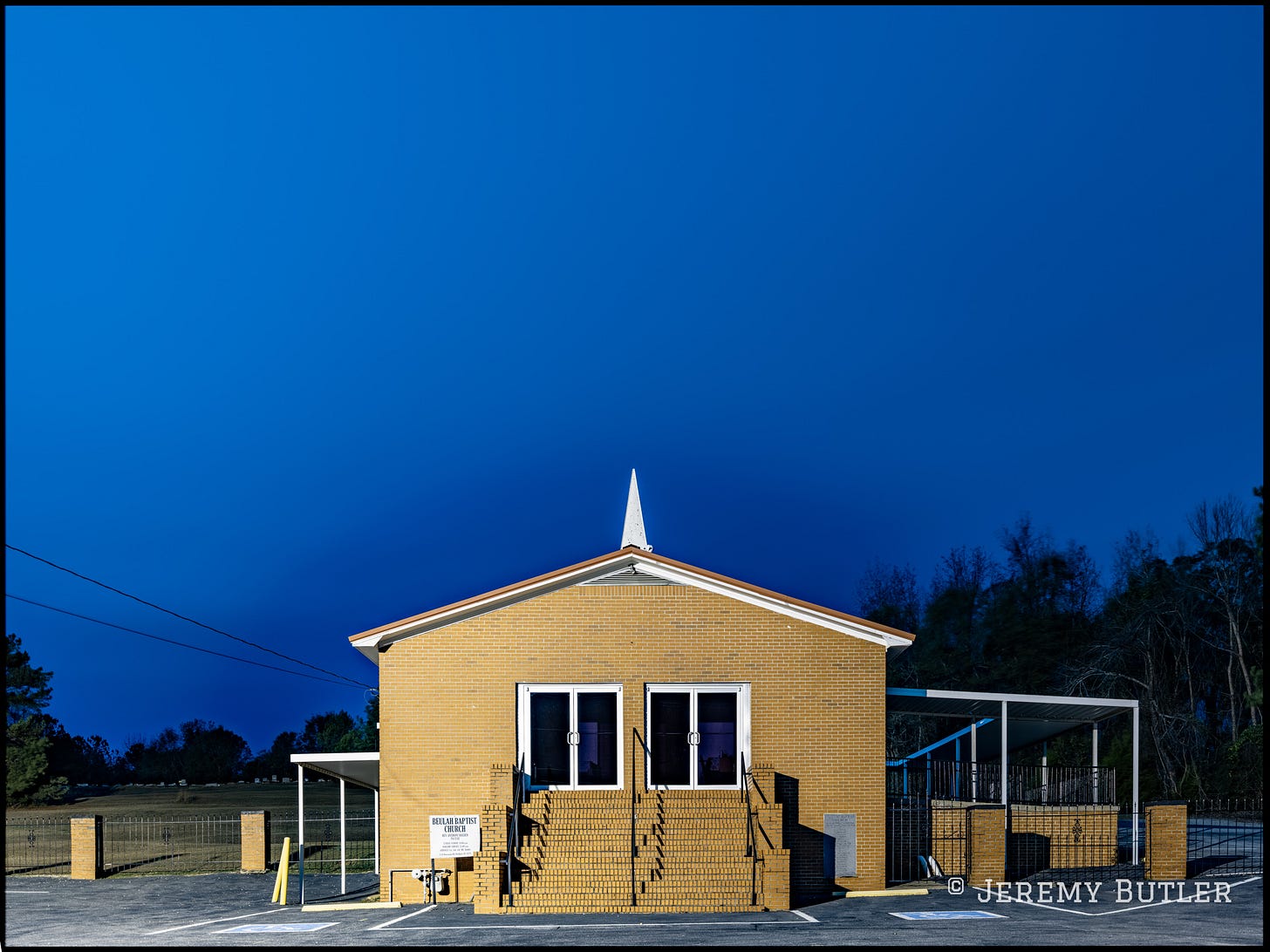 A small yellow brick Baptist church with a white steeple photographed at dusk against a deep blue sky.
