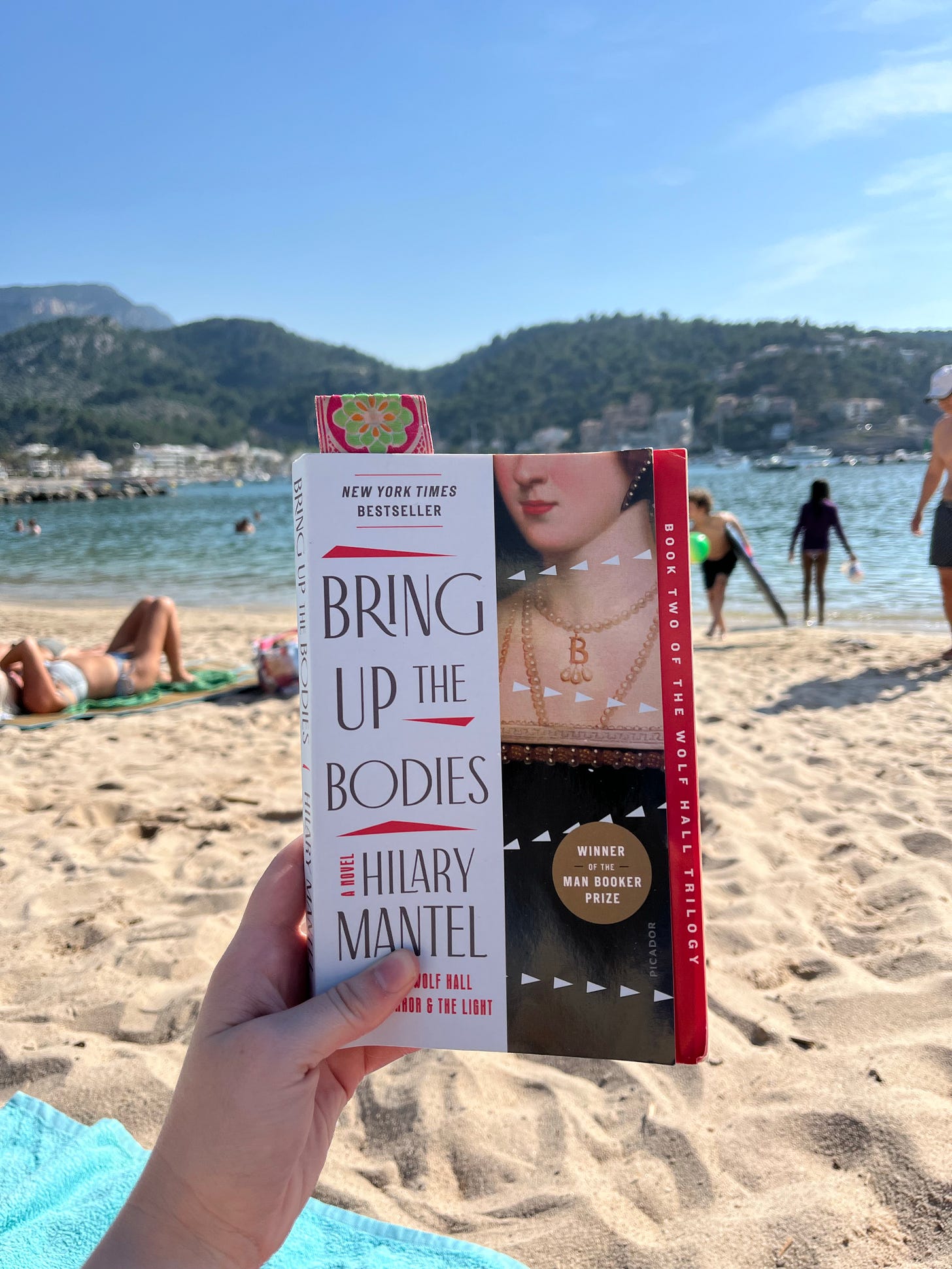 A hand holding up the book Bring Up the Bodies by Hilary Mantel in front of the water on a Mallorcan beach