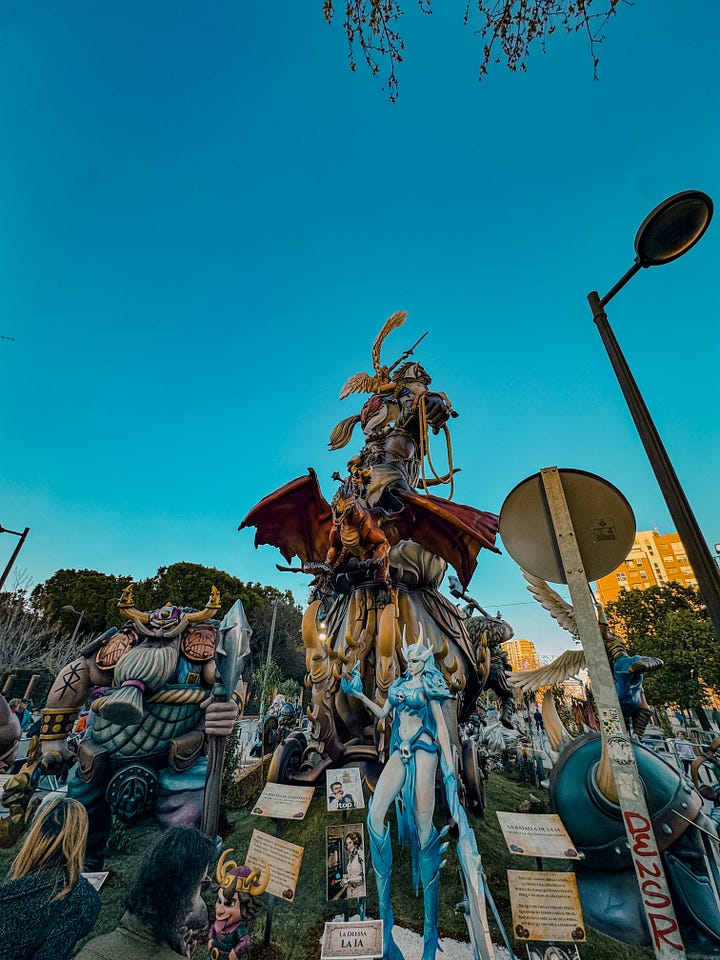A gallery of Fallas monuments in Valencia, Spain — elaborately sculpted figures including a Charlie Chaplin figure holding butterflies, fantasy warriors, and colorful street sculptures, photographed against dramatic cloudy and blue skies in March 2026.