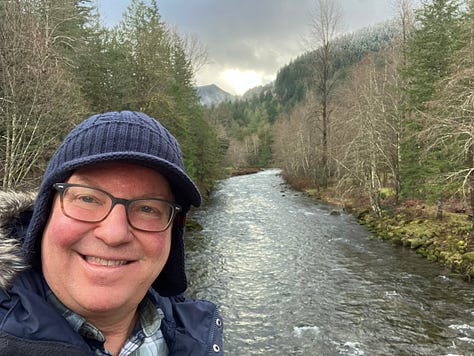 Kacey is wearing a hat and glasses by a river, holding his books by a bookstore, and holding a clipboard at the food distribution center