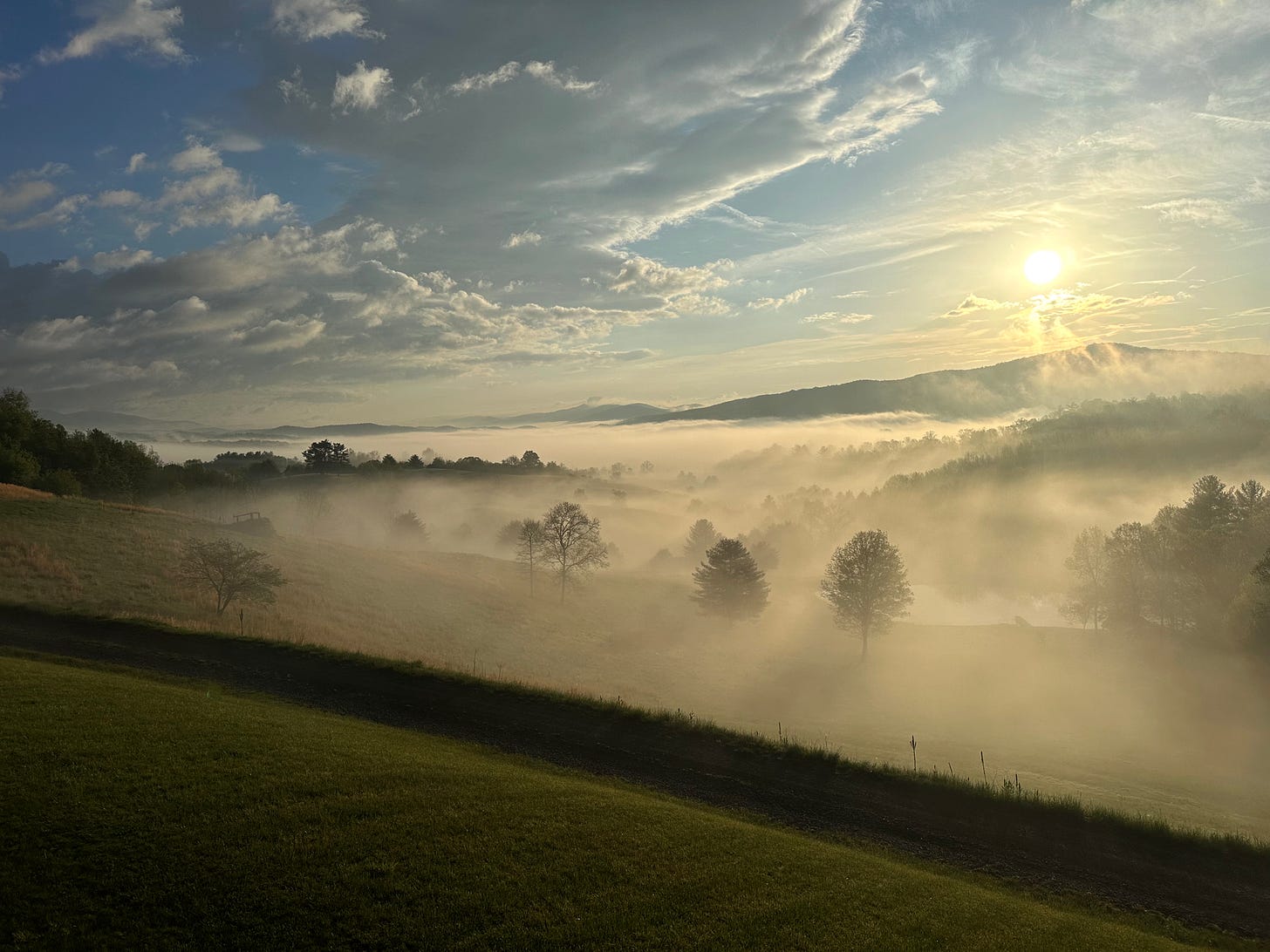 An incredible sunrise over mountains with mist covering them and light green peaking through. Blue and white sky in the distance and a yellow hue over the mist from the sun. 