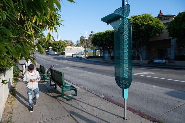 A rectangular, perforated piece of metal is attached to a pole at a bus stop on a sidewalk. The top of the piece of metal is curved, almost at a right angle to the rest of the piece.