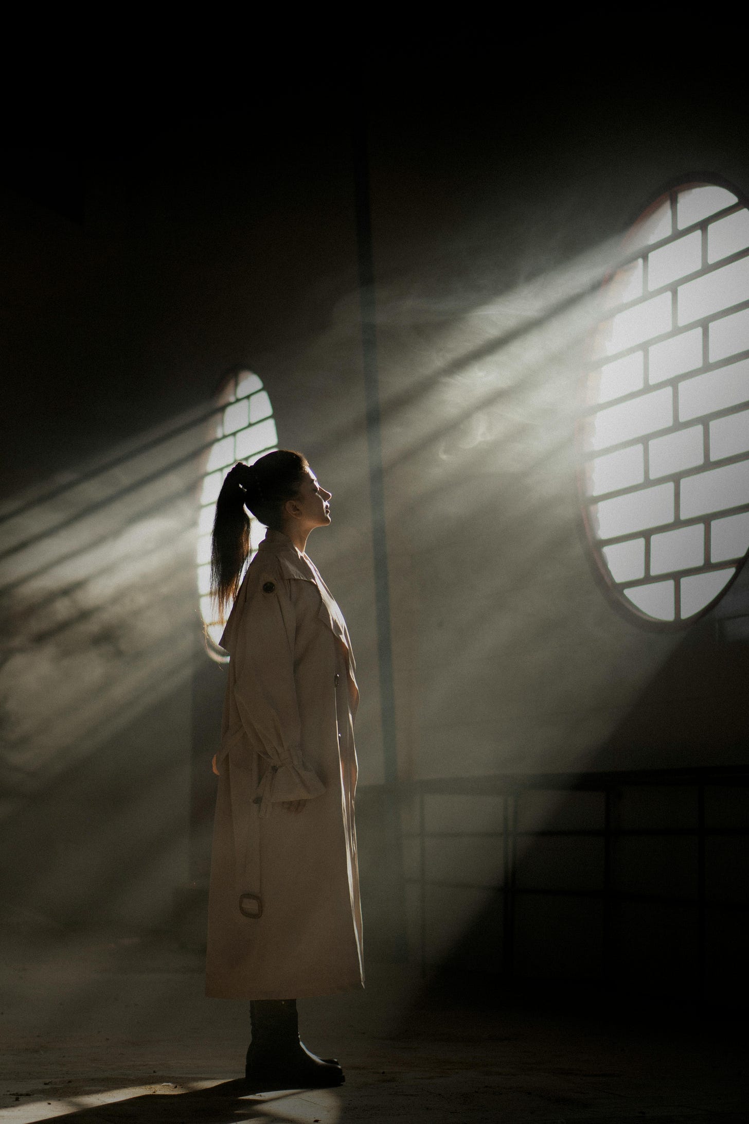 A woman stands in stillness, caught in a shaft of light filtering through a circular window. Dust and shadow fill the air around her, and the scene feels both tender and transcendent.