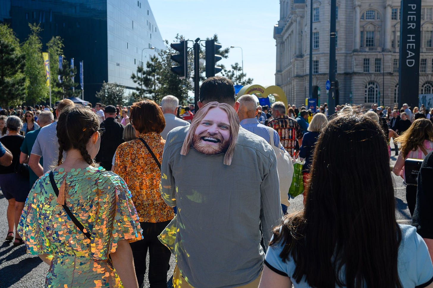A man wears a mask of Sam Ryder smiling on their back as they walk through a crowd.