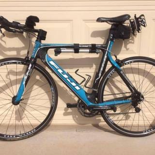 Blue triathlon bike with black aerobars leaning against a beige door. There is a black water bottle holder on the back of the seat, one on the down tube, and one on the seat tube. 