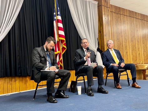The image shows a diverse group of people seated in rows of chairs in a wooden-paneled hall, attentively facing forward. Many attendees are older adults, and some are wearing uniforms or hats indicating military or veteran status. The room is decorated with framed photos, memorabilia, and a disco ball hanging from the ceiling. There are several empty chairs with brochures or program booklets placed on them in the foreground. The atmosphere suggests a formal gathering or community event, likely related to veterans or a commemorative occasion. No specific brands, landmarks, or recognizable public figures are visible.