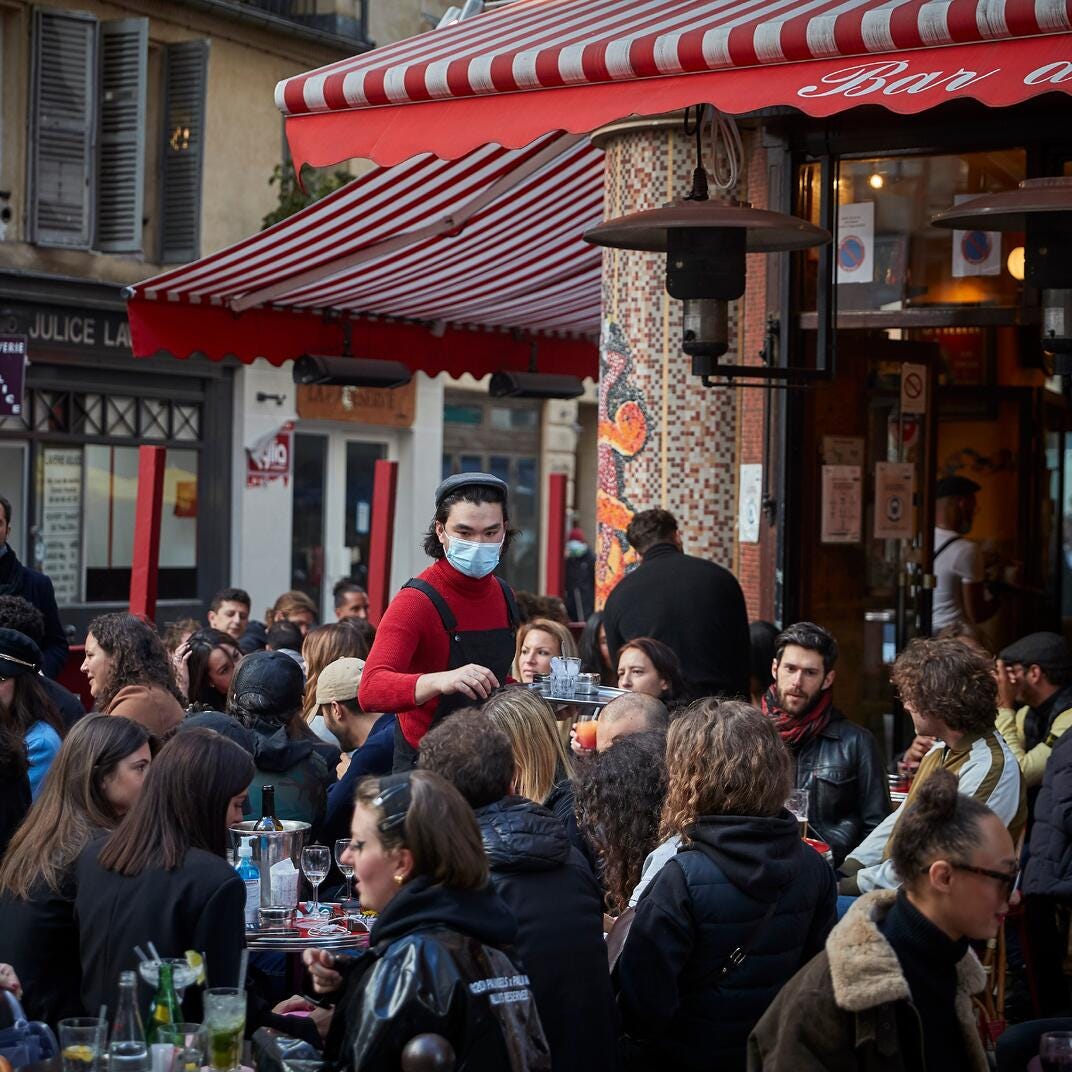 Crowded café/restaurant scene showing a bustling crowd being served by a staff member wearing a mask