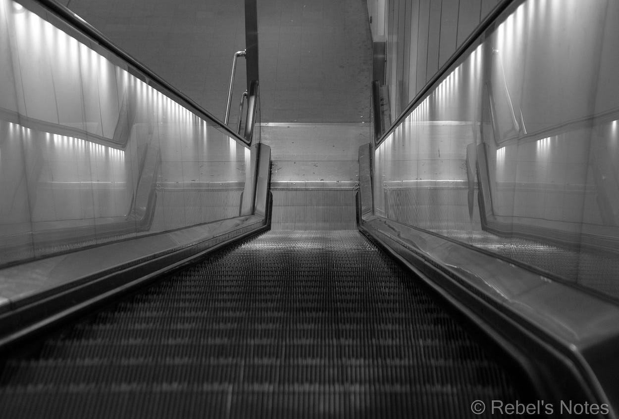 A black and white image of an escalator, where the point of view is from the top.