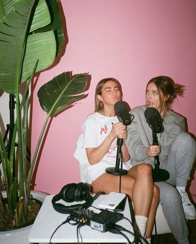 This may contain: two women sitting next to each other in front of a pink wall talking into microphones