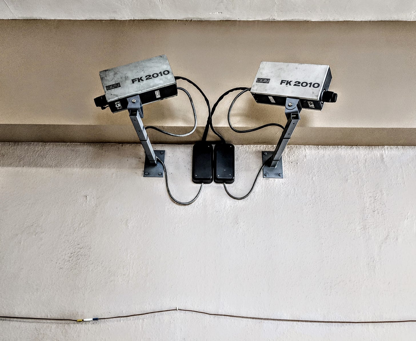 Two outdated security cameras point in opposite directions down a hallway. Text printed on the side of each says "FK 2010." Two outdated security cameras point in opposite directions down a hallway. Text printed on the side of each says "FK 2010."
