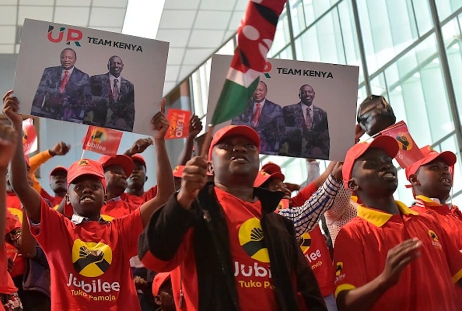 Men in red caps and T-shirts written 'Jubilee Tuko Pamoja' cheering while holding up the Kenyan flag and banners written 'UR Team Kenya' that have two smiling men in suits. Men in red caps and T-shirts written 'Jubilee Tuko Pamoja' cheering while holding up the Kenyan flag and banners written 'UR Team Kenya' that have two smiling men in suits.