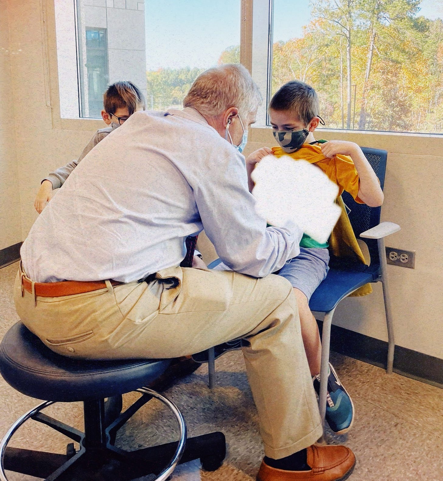 a middle aged male doctor sits on a stool and examines a boy's tummy with his stethoscope. His brother sits next to him.