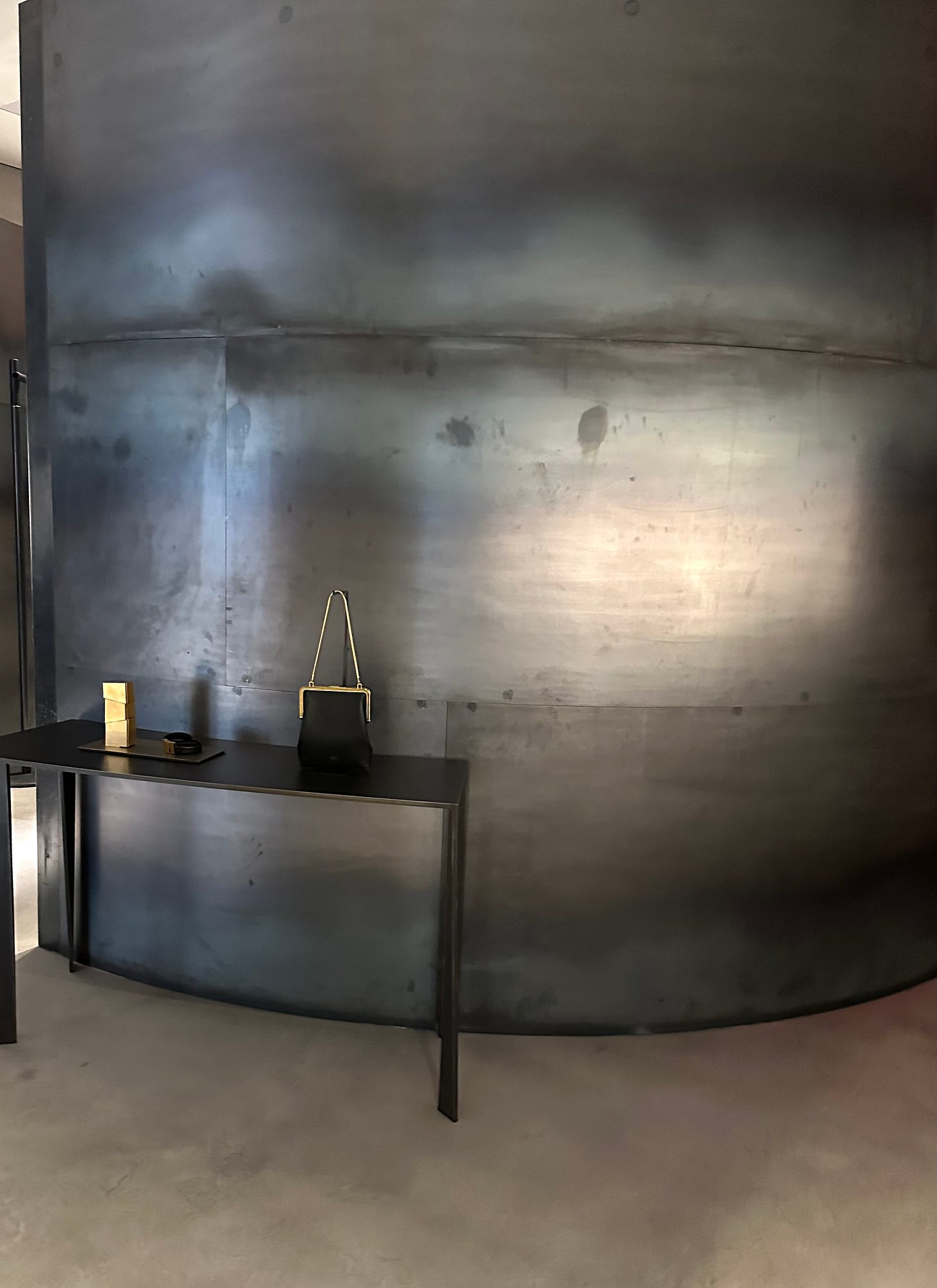 Curved steel wall inside Khaite’s SoHo boutique with a single black top-handle bag on a slim console