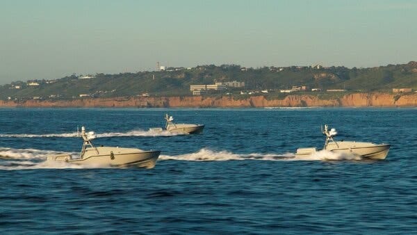 Three driverless boats create waves on the open water backed by land. 