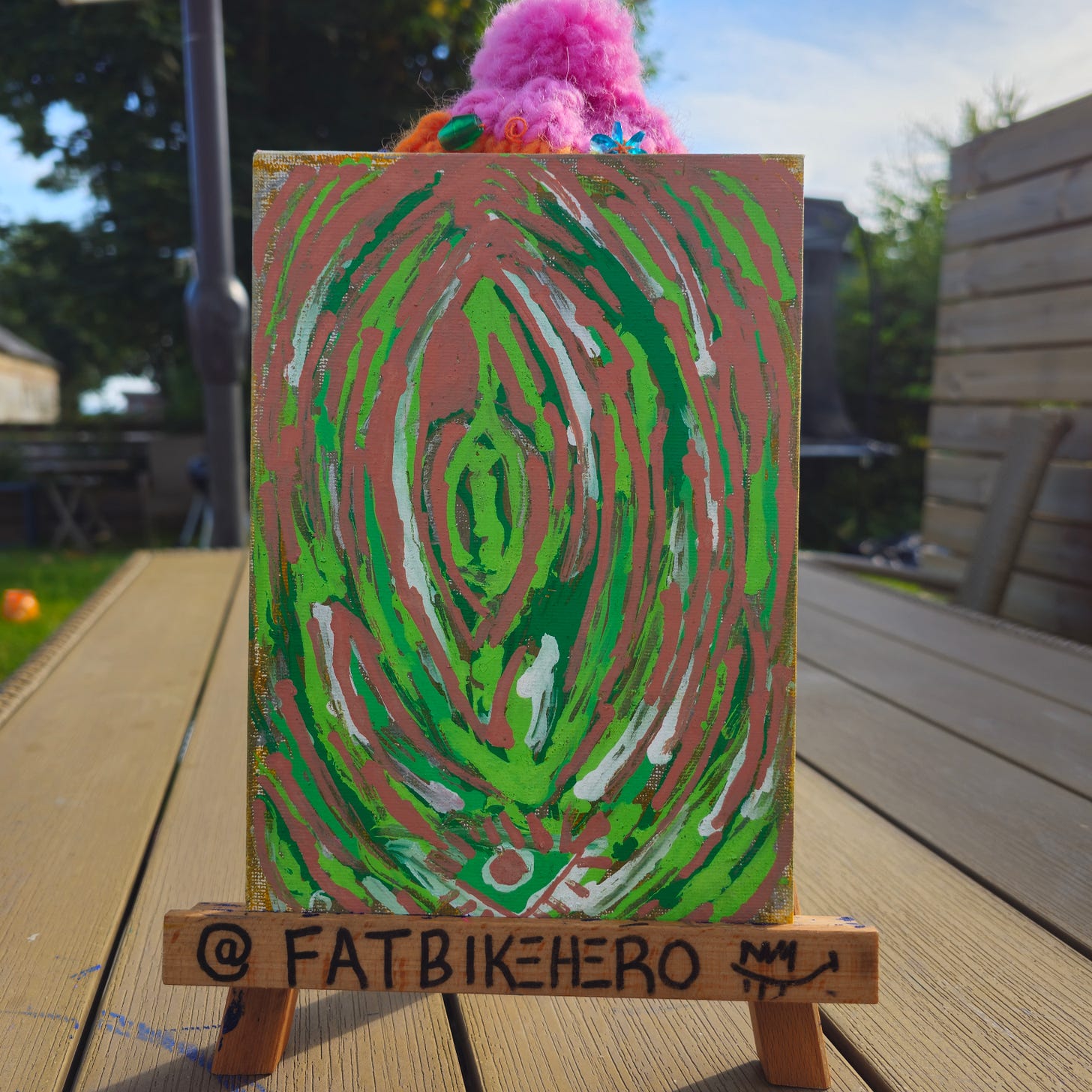 Alt Text: An abstract acrylic painting by FatbikeHero titled "Beautiful Votex." The composition features concentric vertical ovals in neon green, forest green, and terracotta pink with white highlights, resembling a mystical eye or portal. The painting sits on a miniature wooden easel labeled "@FATBIKEHERO" outdoors in natural sunlight. Alt Text: An abstract acrylic painting by FatbikeHero titled "Beautiful Votex." The composition features concentric vertical ovals in neon green, forest green, and terracotta pink with white highlights, resembling a mystical eye or portal. The painting sits on a miniature wooden easel labeled "@FATBIKEHERO" outdoors in natural sunlight.