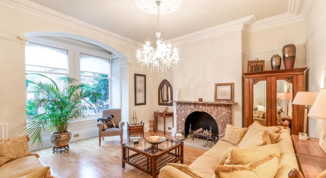 A cream living room with an arched bay window, a hanging chandelier, a wooden coffee table, and light yellow seats