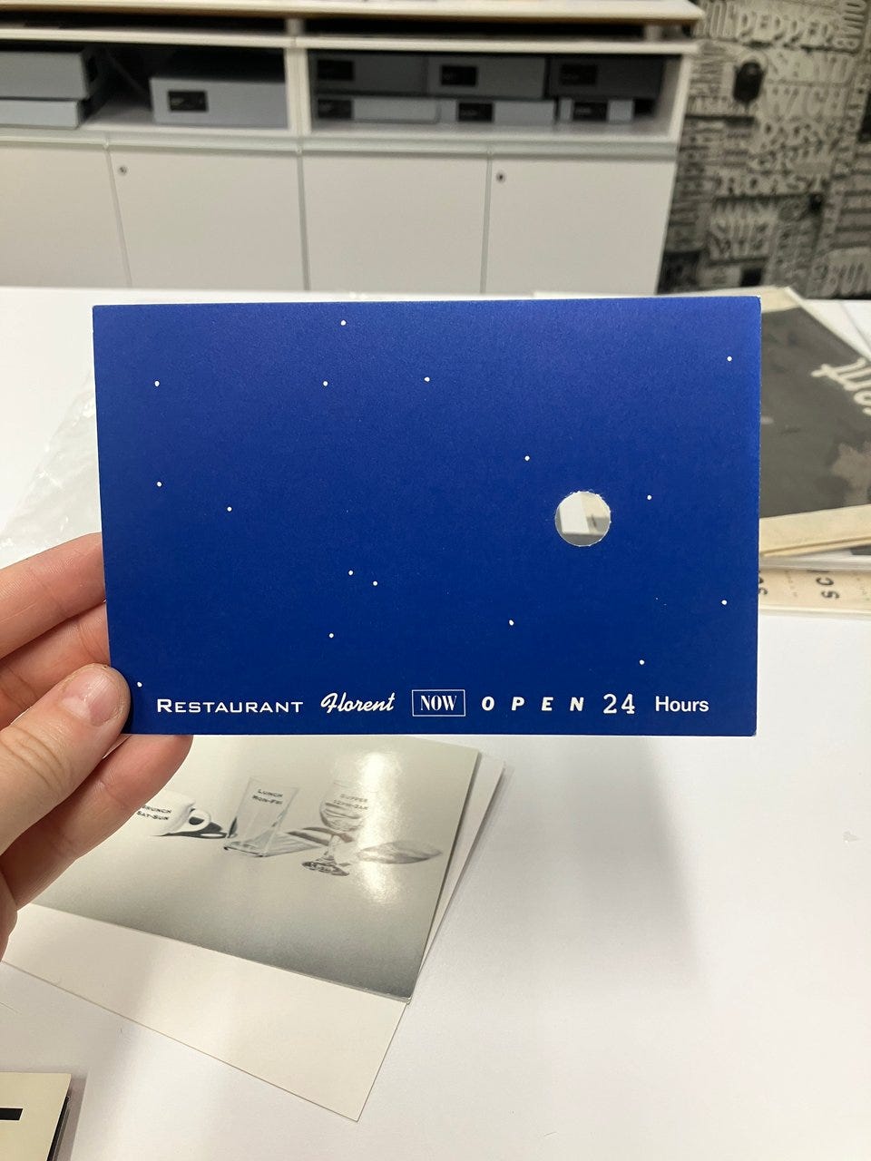 blue postcard showing a starry night with a hole cut out. "restaurant florent now open 24 hours" written underneath in all diferent fonts