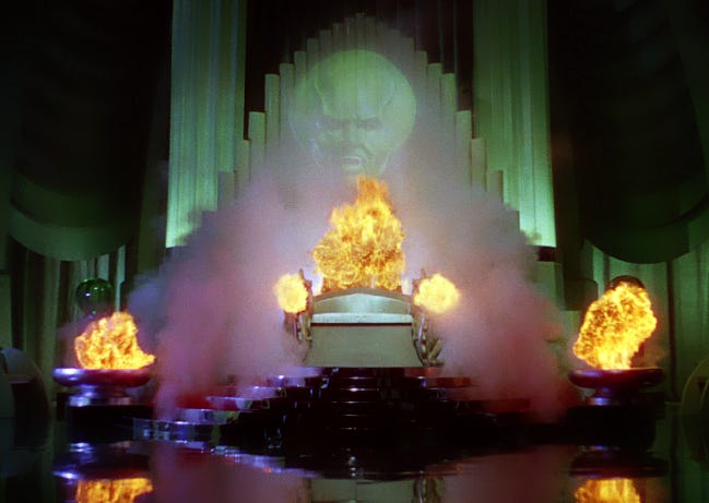 Image of "The Wizard of Oz" from the original film, fire bellows from cauldrons as the green-faced wizard's head floats in a plume of purple smoke above a set of staris