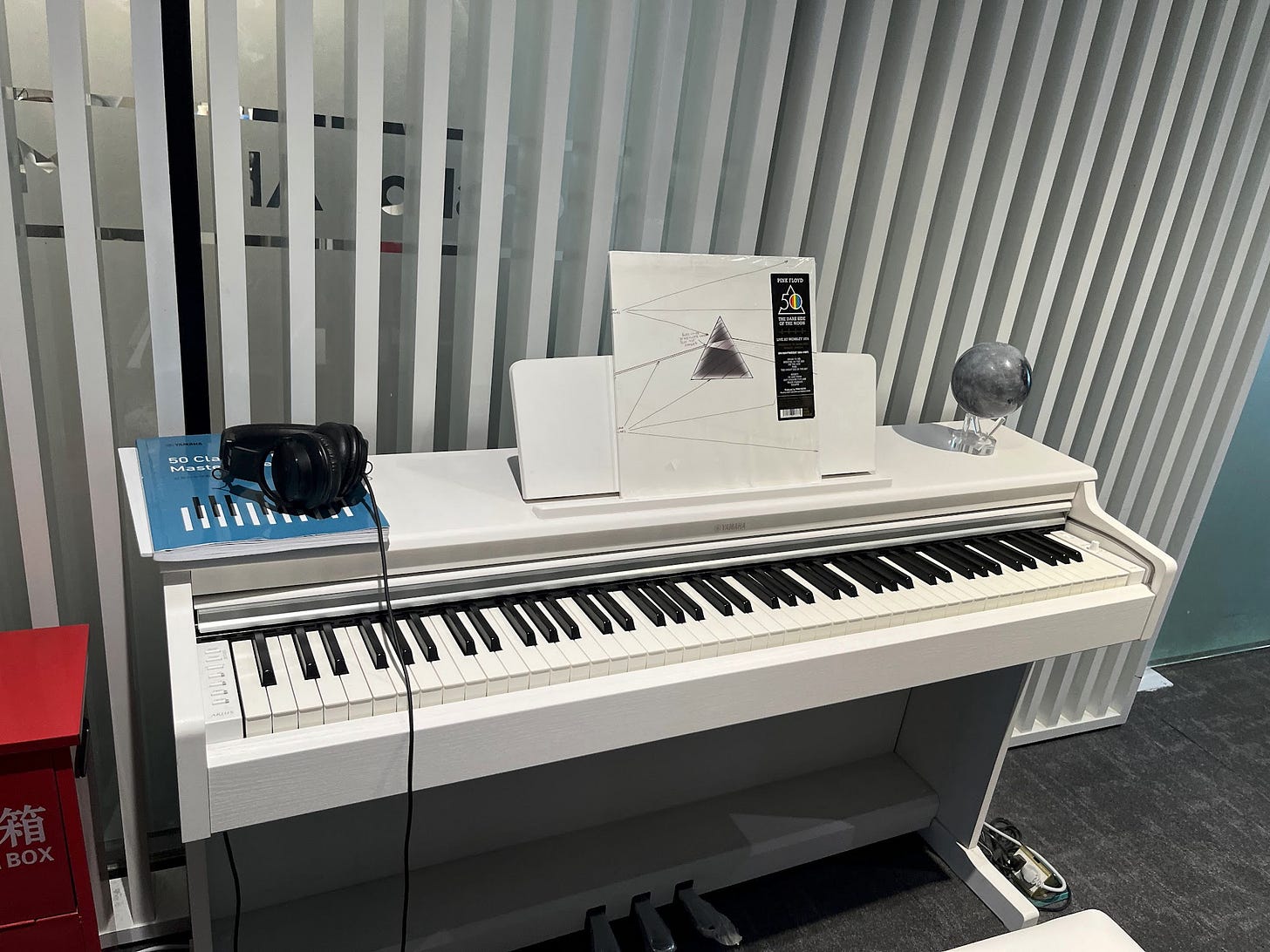 A white Yamaha upright digital piano in the lobby of the Moonshot AI office, with a vinyl copy of Pink Floyd's "The Dark Side of the Moon" 50th anniversary edition displayed on the music stand.