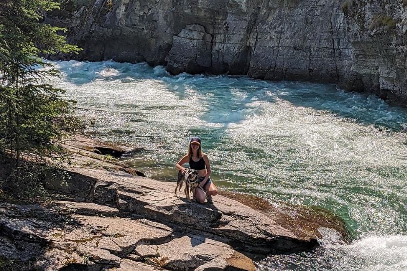 A young woman poses with a blue heeler on a rock at the edge of a blue river