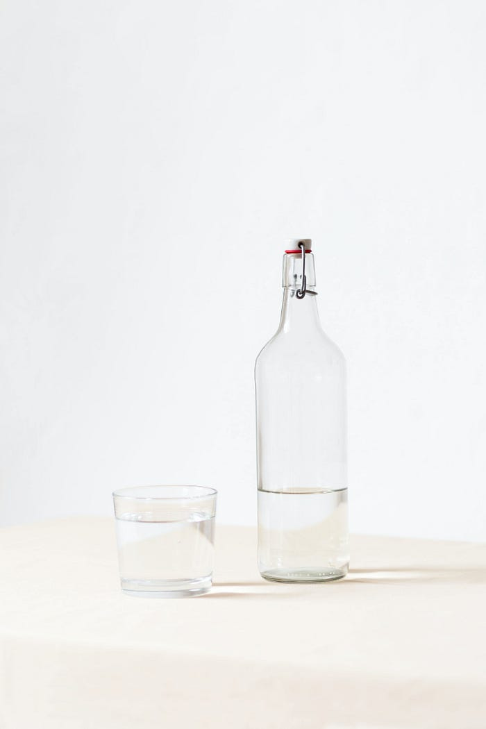 A cup full of water with a near-empty glass bottle beside it. A cup full of water with a near-empty glass bottle beside it.