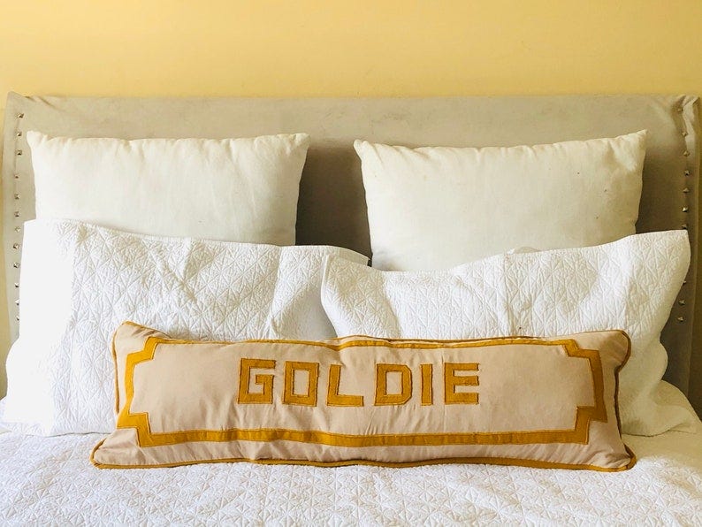 May include: A beige pillow with a yellow border and the word 'GOLDIE' embroidered in yellow on the front. The pillow is on a bed with white bedding and two white pillows.