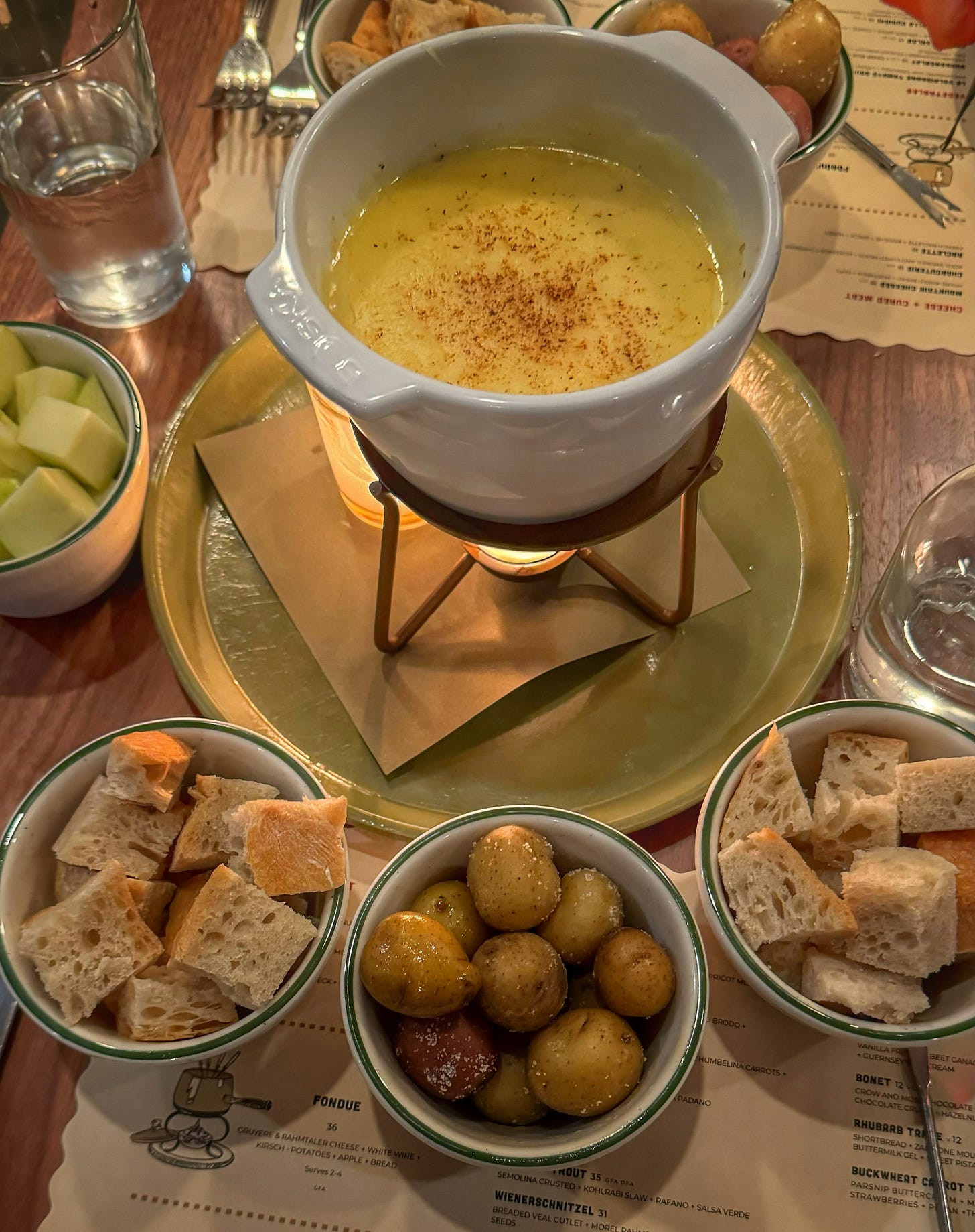 A small fondue pot full of melted cheese, being warmed by a tealight. Surrounding the fondue pot are small bowls with chunks of bread and apple and baby potatoes with large flakes of salt on them. 