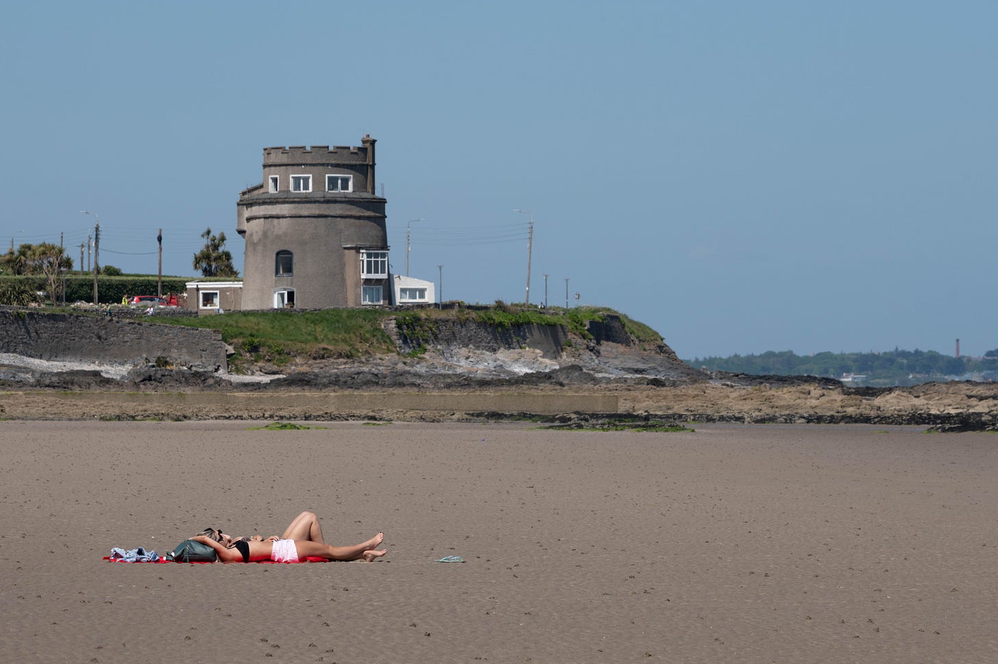 A person sunbathes on a wide sandy beach beneath a Martello tower, the stone structure standing against a clear blue sky A person sunbathes on a wide sandy beach beneath a Martello tower, the stone structure standing against a clear blue sky