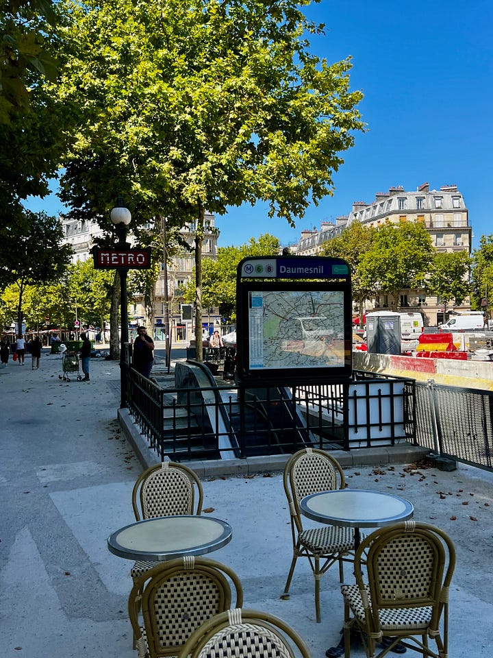 (emptEmpty café chairs arranged outside the Daumesnil metro station entrance on a quiet afternoon, with trees casting shadows on the pavement. Row of shuttered storefronts along an empty Parisian street, with closed metal security gates pulled down over shop windows.