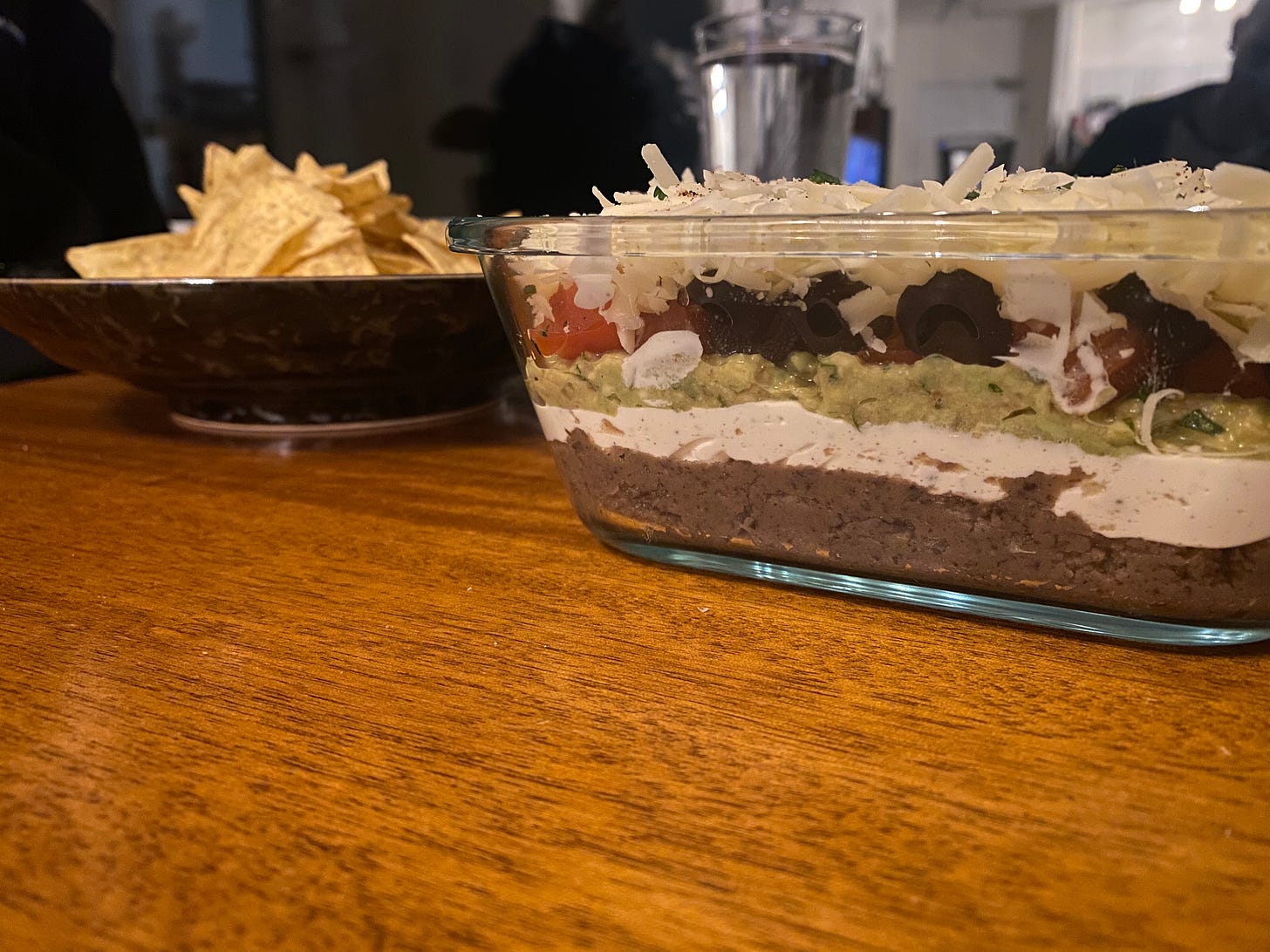 A glass dish of seven layer dip, photo taken at eye level so you can see all the layers. Behind it is a shallow bowl of tortilla chips.
