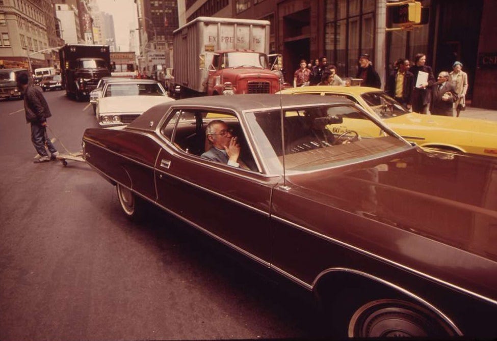 An enormous 2-door Mercury in Manhattan traffic in 1973. A man in the passenger seat smokes a cigarette, the window down. Behind that car is heavy car and truck traffic and a jaywalking man pulling an empty piano dolly.    