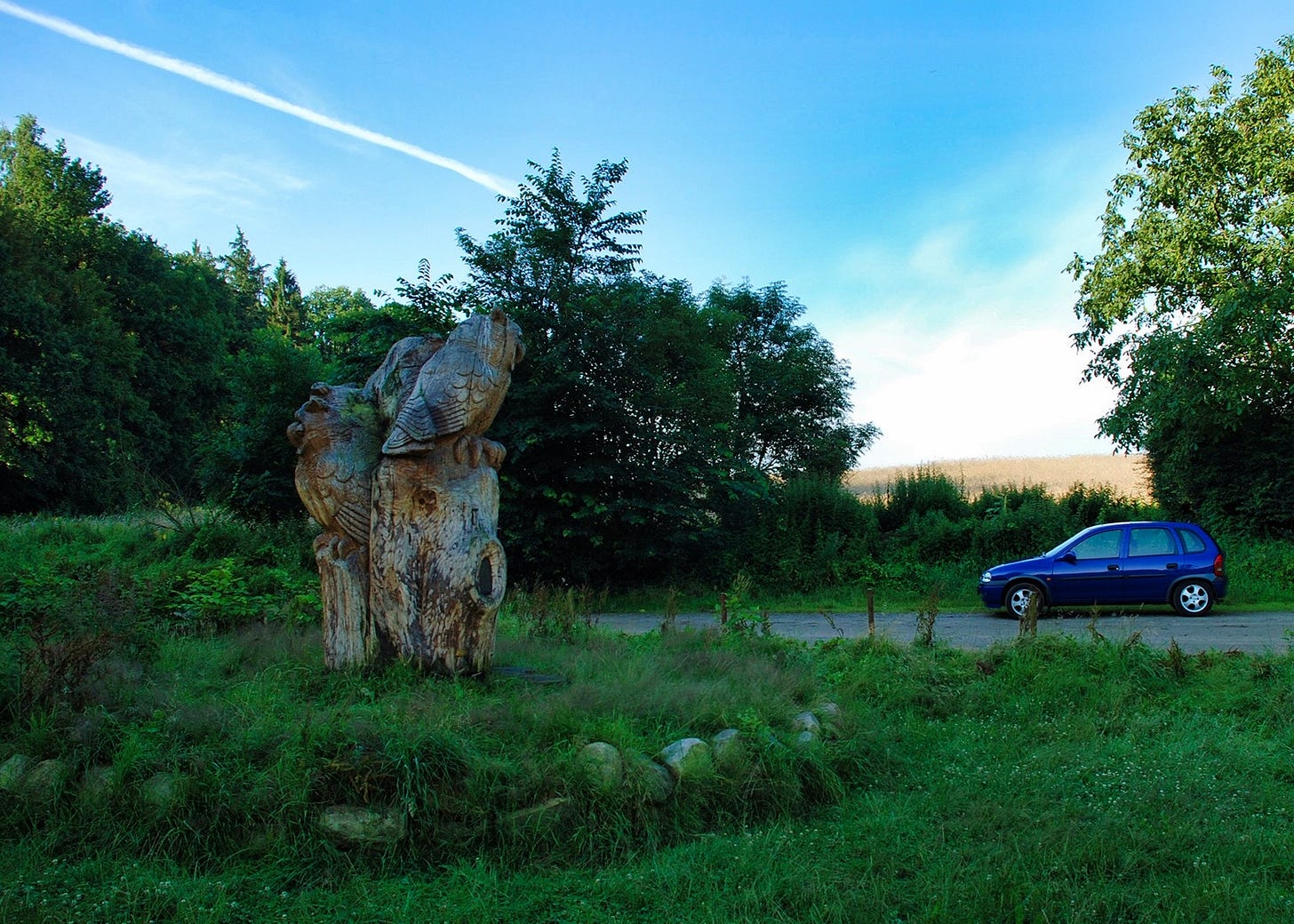 Große Holzskulptur auf einer Wiese am Waldrand, aus einem Baumstamm geschnitzt und zwei Eulen darstellend. Rechts daneben ein blauer Kleinwagen auf einem Schotterweg. Im Hintergrund dichte Bäume und ein bewaldeter Hügel unter blauem Himmel mit einem Kondensstreifen. Große Holzskulptur auf einer Wiese am Waldrand, aus einem Baumstamm geschnitzt und zwei Eulen darstellend. Rechts daneben ein blauer Kleinwagen auf einem Schotterweg. Im Hintergrund dichte Bäume und ein bewaldeter Hügel unter blauem Himmel mit einem Kondensstreifen.